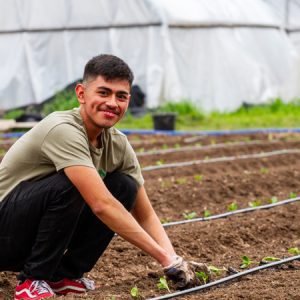 An Urban Roots youth in a green shirt and red shoes works in a field planting sprouts, smiling.