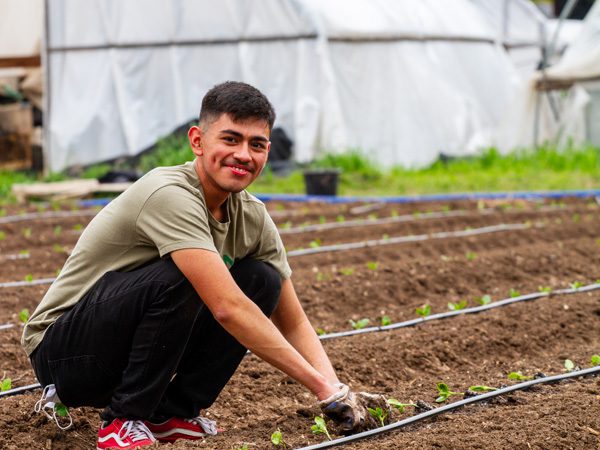 An Urban Roots youth in a green shirt and red shoes works in a field planting sprouts, smiling.